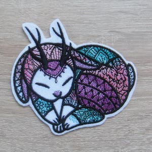 May include: A whimsical patch featuring a stylized rabbit with deer-like antlers. The patch is primarily white with intricate embroidery in shades of purple, teal, and pink. The rabbit is in a resting pose, with closed eyes and detailed patterns.