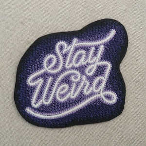 Op de afbeelding: Paarse en witte geborduurde strijkpatch met de tekst "Stay Weird" in een cursief schrift.