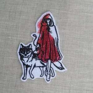 以下が含まれることがあります： 赤いフード付きのマントを着た女の子が狼と歩いている刺繍パッチ。パッチは白地に黒のアウトラインと赤いステッチで仕上げられています。