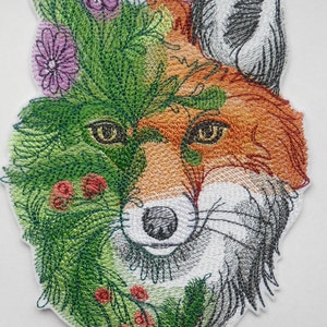 Peut inclure: Patch brodé représentant le visage d'un renard avec de la végétation verte, des baies rouges et des fleurs violettes. La fourrure du renard est orange, blanche et grise.