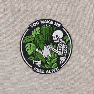 Könnte beinhalten: Schwarzer und weißer gestickter Aufnäher mit einem Skelett, das Pflanzen gießt. Der Aufnäher ist rund und hat den Text "You Make Me Feel Alive" am Rand.
