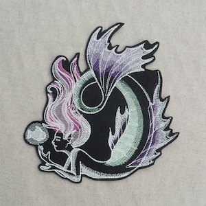 May include: Embroidered patch featuring a mermaid with pink hair holding a pearl, with a green and purple tail against a black background.