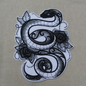 May include: Black and white embroidered patch featuring a snake wrapped around roses. The snake is curled up and has a detailed design. The roses are stylised and have a delicate look.