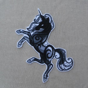 May include: Black embroidered unicorn patch featuring intricate lace-like details. The unicorn is standing on its hind legs with its head turned to the side. The patch is ideal for adding a touch of whimsy to clothing, bags, or other accessories.