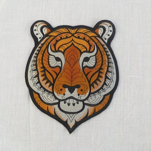 May include: An embroidered patch featuring a detailed tiger's face in orange, black and white. The tiger's face is stylised with intricate patterns and designs.