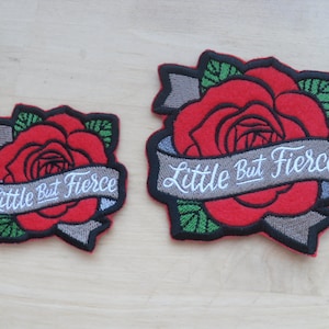May include: Two embroidered iron-on patches featuring a red rose with green leaves and a grey banner with the text "Little But Fierce" in white lettering. The larger patch is approximately 7.5 cm in diameter, while the smaller patch is approximately 5 cm in diameter.