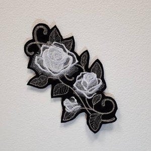 May include: White embroidered roses on a black fabric patch. The roses are arranged in a cluster with leaves and stems.