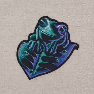 Könnte beinhalten: Ein gestickter Aufnäher mit einem grünen Frosch, der auf einem lila und grünen Blatt sitzt. Der Frosch hat große, schwarze Augen und ein breites Lächeln.