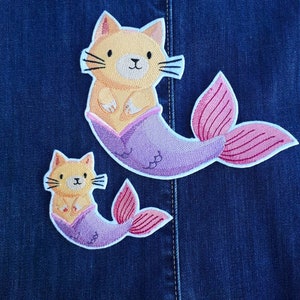 Op de afbeelding: Twee geborduurde strijkpatches van oranje katten met paarse en roze zeemeerminstaarten. De grotere patch is bovenaan en de kleinere patch is onderaan.