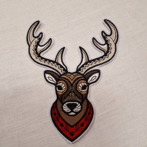 May include: An embroidered patch featuring a deer's head with intricate brown and white detailing. The deer is wearing a red and white patterned bandana.