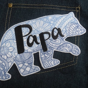Può includere: Un'applicazione a forma di orso con un motivo blu e bianco e la parola "Papà" ricamata in filo nero.