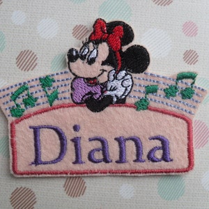 Pode incluir: Patch bordado com Minnie Mouse com laço vermelho, vestido roxo e luvas brancas. O patch tem um fundo rosa claro com o nome "Diana" em letras roxas. Notas musicais verdes estão acima do nome.