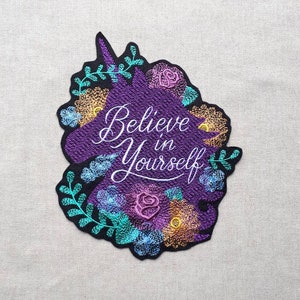 May include: Purple embroidered iron-on patch with a unicorn silhouette surrounded by flowers and leaves. The text "Believe in Yourself" is embroidered in white on the unicorn.