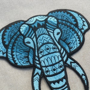 May include: Blue embroidered elephant head patch with intricate geometric patterns. The patch is made of fabric and has a black outline.
