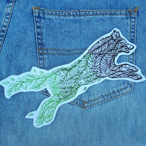 Może przedstawiać: Naszywka z haftowanym wizerunkiem skaczącego wilka, którego ciało tworzą liście, w kolorze zielonym i czarnym, na tle niebieskiego dżinsu.