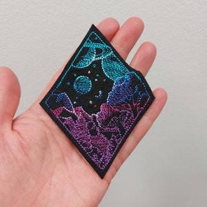 以下が含まれることがあります： 三日月と星が輝く、紫、青、ターコイズブルーの山々が描かれた、黒い菱形の刺繍パッチ。