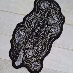 May include: Embroidered patch featuring a detailed tiger design in shades of gray and white, set against a black background. The patch has a unique, irregular shape and includes decorative circular elements.