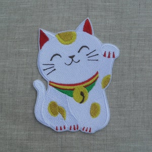 Op de afbeelding: Witte geborduurde patch van een glimlachende, zittende kat met een opgeheven poot. De kat heeft gele vlekken en een groene halsband met een gouden bel. De kat is een traditionele Japanse Maneki-Neko, of "wenkende kat", een geluksbrenger.