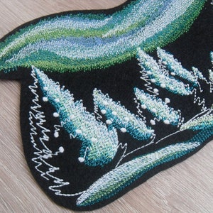 Pode incluir: Etiqueta bordada com fundo de feltro preto que apresenta um design de aurora boreal verde e azul sobre uma fileira de árvores brancas e verdes.