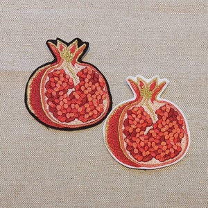 Pode incluir: Dois patches bordados para passar a ferro de granadas vermelhas com detalhes dourados. As granadas estão abertas e mostram as sementes no interior.