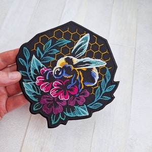 May include: A black embroidered patch featuring a bee flying over a honeycomb and flowers. The bee is yellow, black, and blue. The flowers are pink, purple, and green.