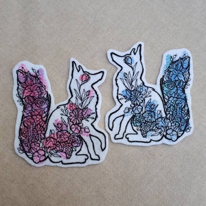 以下が含まれることがあります： 花の模様でいっぱいのキツネのシルエットが刺繍された2つのパッチ。1つのパッチはピンクとパープル、もう1つはブルーとグリーンです。