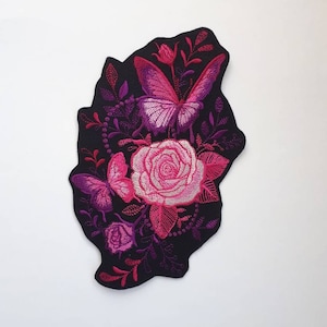 Può includere: Toppa ricamata con una rosa rosa e viola con due farfalle rosa e viola su sfondo nero.