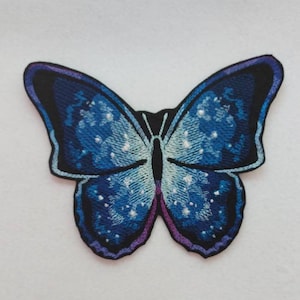 Può includere: Una toppa ricamata a forma di farfalla blu e viola con un motivo bianco e blu. La farfalla è su uno sfondo bianco.