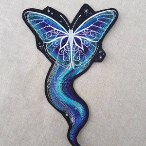 Könnte beinhalten: Ein gestickter Schmetterlingspatch in Blautönen und Lila mit weißen Details und einem wirbelnden Design. Der Schmetterling fliegt mit einem blauen und lilafarbenen Schweif nach oben.