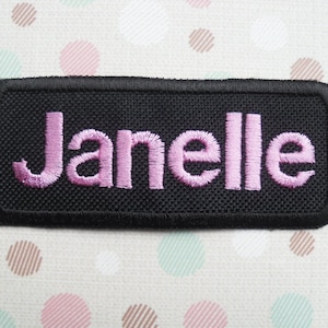 Può includere: Toppa rettangolare nera con il nome "Janelle" ricamato in filo rosa.