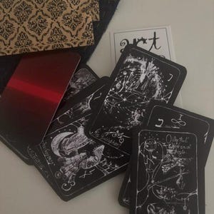 May include: A collection of black tarot cards with white line art illustrations and text, including cards labelled The Empress, The Hierophant, and O the fool! Where we all begin. A metallic red card and a tan envelope with a black damask pattern are present.