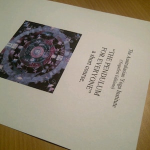 May include: A white paper with text from The Australasian Yoga Institute, including "THE PENDULUM FOR EVERYONE" and a short course description. A colorful circular graphic is at the bottom of the page. The paper is on a wooden surface.