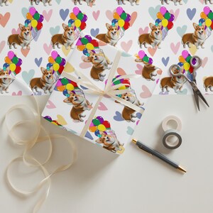 May include: Wrapping paper with a repeating pattern of corgi dogs holding balloons, and hearts. A wrapped gift box with a cream ribbon is in the centre. Scissors, tape, and a pen are also visible.