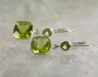 May include: A pair of silver dangle earrings with green gemstone accents. The earrings feature two green gemstones, one larger and one smaller, on each earring.