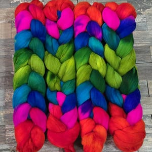 May include: Four colorful braids of dyed wool in vibrant hues of red, pink, blue, and green. The wool is arranged in a thick, textured pattern, showcasing the blend of colors. The braids are displayed against a neutral background.