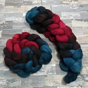 May include: Two braids of colorful wool roving. One is a long braid, and the other is a circular braid. The colors transition from teal to dark brown to red. The wool is soft and fluffy, perfect for fiber art projects.