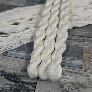 May include: Close-up of off-white yarn skeins. The yarn is tightly twisted, with a soft, slightly shiny texture. The background is a weathered wooden surface with various shades of gray and brown.