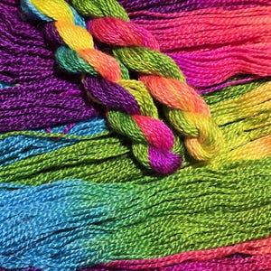 May include: Close-up of colorful embroidery floss in shades of purple, blue, green, yellow, orange, and pink. The floss is arranged in bundles and a braided strand, showcasing the vibrant color variations.