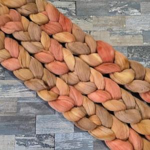 May include: Close-up of a thick, braided yarn in shades of peach, tan, and brown. The yarn is arranged diagonally against a gray, textured background, creating a visually appealing pattern. The texture of the yarn is soft and inviting.