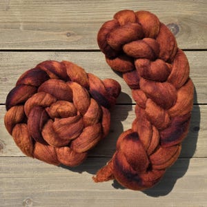 May include: Two skeins of hand-dyed wool roving in shades of orange and brown. The wool is arranged in a braided pattern, with darker hues at the center and edges. The background is a light-colored wooden surface.