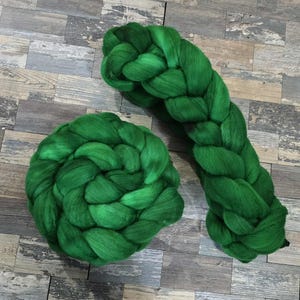 May include: Two bundles of vibrant green wool roving. One is coiled, the other is a long, thick braid. The wool has a soft, fluffy texture, and the color is a rich, saturated green. The background is a wooden surface.