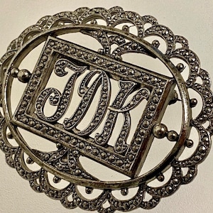 May include: A silver-toned brooch with a decorative oval frame and a rectangular center piece featuring the initials "J.K." in a stylized font.
