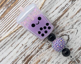 May include: A cute, purple boba tea badge reel with a smiling face and black boba pearls. The reel features a beaded design with a large, sparkly purple bead and black beads. A clear plastic clip and a small, matching purple charm complete the design.