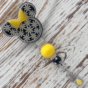 May include: A daisy-themed badge reel featuring a Mickey Mouse silhouette with a yellow glitter bow. The black and white design is accented with a yellow bead and a black and white polka dot bead.