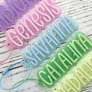 May include: Five colorful name tags with the names Abigail, Genesis, Savanna, Catalina, and Madaline. Each tag is a different color, including purple, pink, blue, green, and yellow-green, with glitter accents and a matching wire loop.