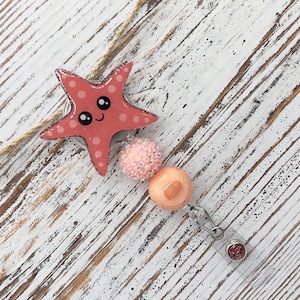 May include: A badge reel featuring a coral-colored starfish with white polka dots and a smiling face. The reel includes pink and peach-colored beads and a glittery accent on the clip. The background is a white-washed wooden surface.