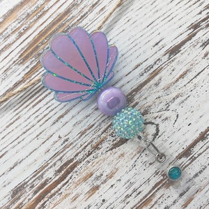 May include: A mermaid-themed retractable badge holder with a seashell design. The badge reel features a lavender seashell, a large purple bead, and a cluster of light blue crystal beads. The reel has a clear plastic base and a silver clip.