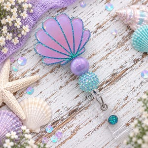 May include: A badge reel featuring a light purple seashell charm with teal glitter accents, attached to a strand of beads including a smooth purple sphere and a textured teal crystal bead, ending in a clear plastic strap with a small teal button.