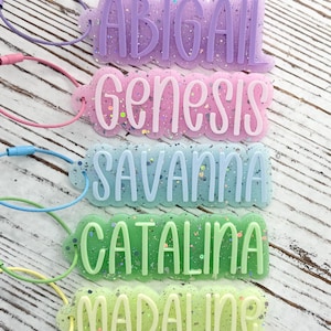 May include: Five rectangular acrylic luggage tags with scalloped edges and glitter accents. Each tag features a raised name in a different color: purple ABIGAIL, pink Genesis, light blue SAVANNA, green CATALINA, and light green MADALINE.