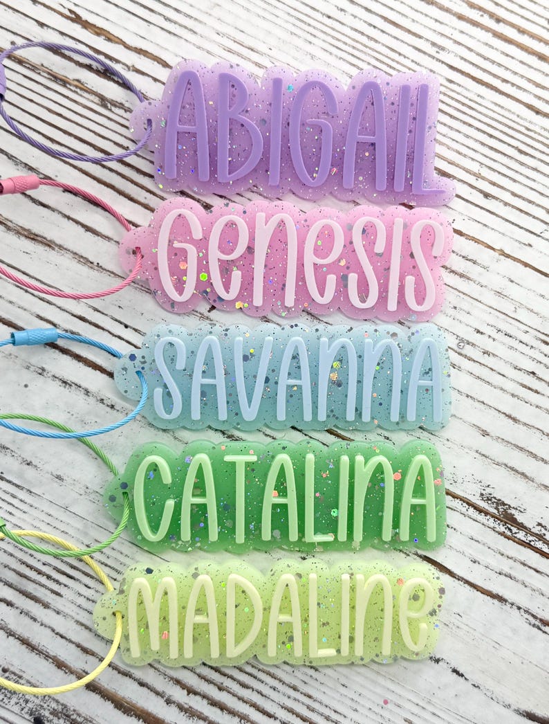 May include: Five rectangular acrylic luggage tags with scalloped edges and glitter accents. Each tag features a raised name in a different color: purple ABIGAIL, pink Genesis, light blue SAVANNA, green CATALINA, and light green MADALINE.